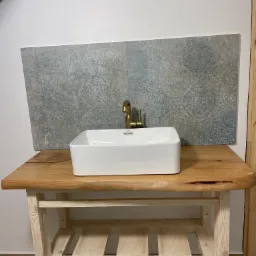 Biała, prostokątna umywalka nablatowa z mosiężną baterią, osadzona na blacie z litego drewna o nieregularnych krawędziach, z półką pod spodem. Na ścianie płytki w odcieniach szarości z subtelnym...