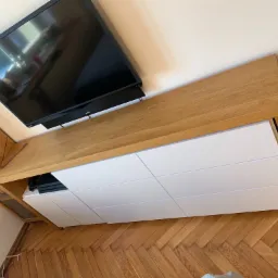 Nowoczesna szafka RTV z blatem z jasnego drewna i białymi frontami, zawieszona pod telewizorem na ścianie, widoczna na tle parkietu ułożonego w jodełkę.