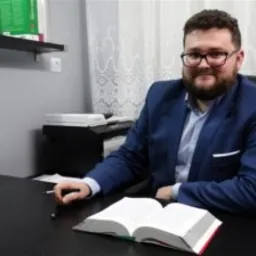 Mężczyzna w okularach i niebieskiej marynarce siedzi za biurkiem z otwartą książką, w tle drukarka i zasłony.