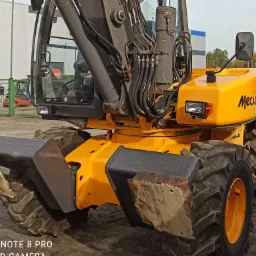 Żółta koparka Mecalac MTX z łyżką, lekko ubrudzona ziemią, na placu budowy z widocznym w tle budynkiem magazynowym i wózkiem widłowym.