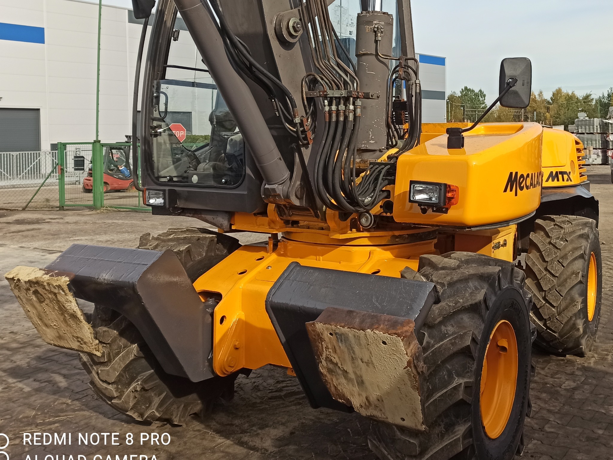 Żółta koparka Mecalac MTX z łyżką, lekko ubrudzona ziemią, na placu budowy z widocznym w tle budynkiem magazynowym i wózkiem widłowym.