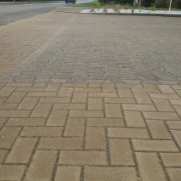 Fragment nawierzchni z kostki brukowej ułożonej w jodełkę, widoczna różnica w czystości między umytą a nieumytą częścią, w tle fragment samochodu i ulicy.