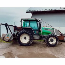 Zielono-szary traktor Valtra z osprzętem do robót ziemnych, zaparkowany na mokrym terenie w Myślenicach, na tle budynku z rynnami ozdobionymi motywem koni.
