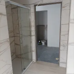 Wykończona łazienka z kabiną prysznicową i marmurowymi płytkami na ścianach, widok na sąsiednie pomieszczenie z szarymi płytkami podłogowymi i wiadrami po farbie.