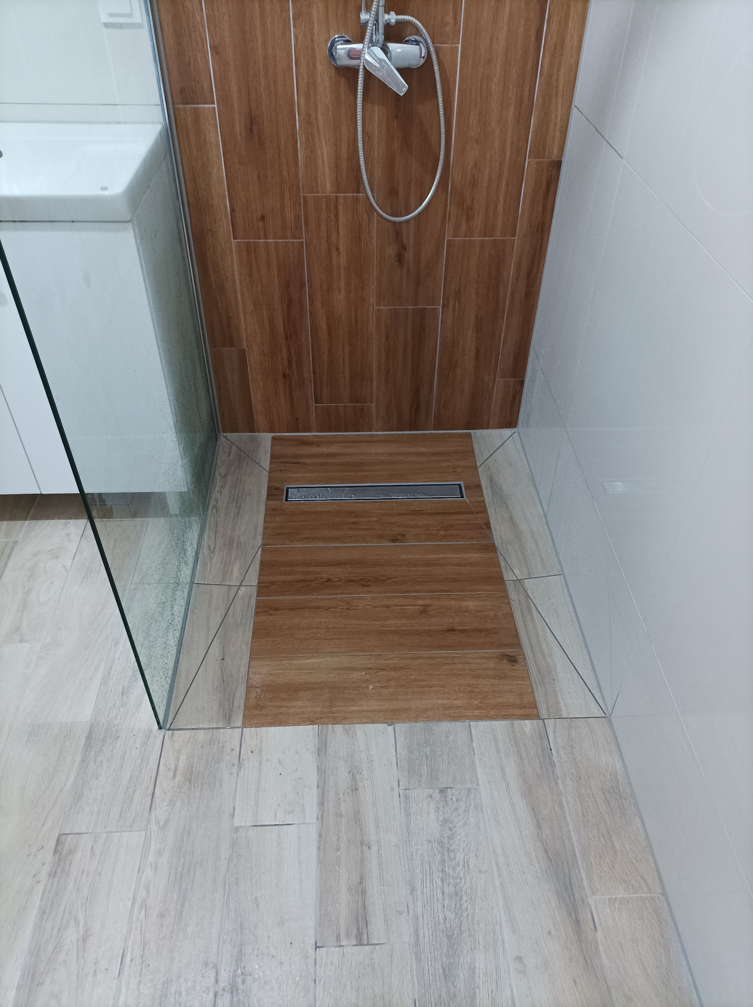 Wykończona kabina prysznicowa z odpływem liniowym, brązowe i beżowe płytki imitujące drewno, widoczna bateria prysznicowa i fragment szklanej ściany kabiny.