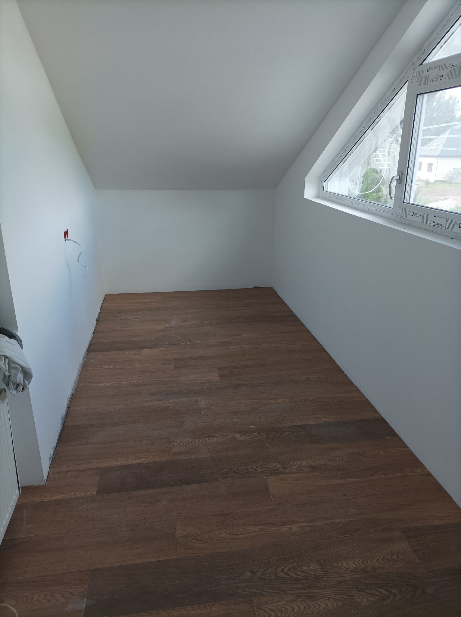 Podłoga z paneli drewnopodobnych w pokoju na poddaszu z oknem dachowym, widoczne niedokończone wykończenia przy ścianach i wystające kable elektryczne.
