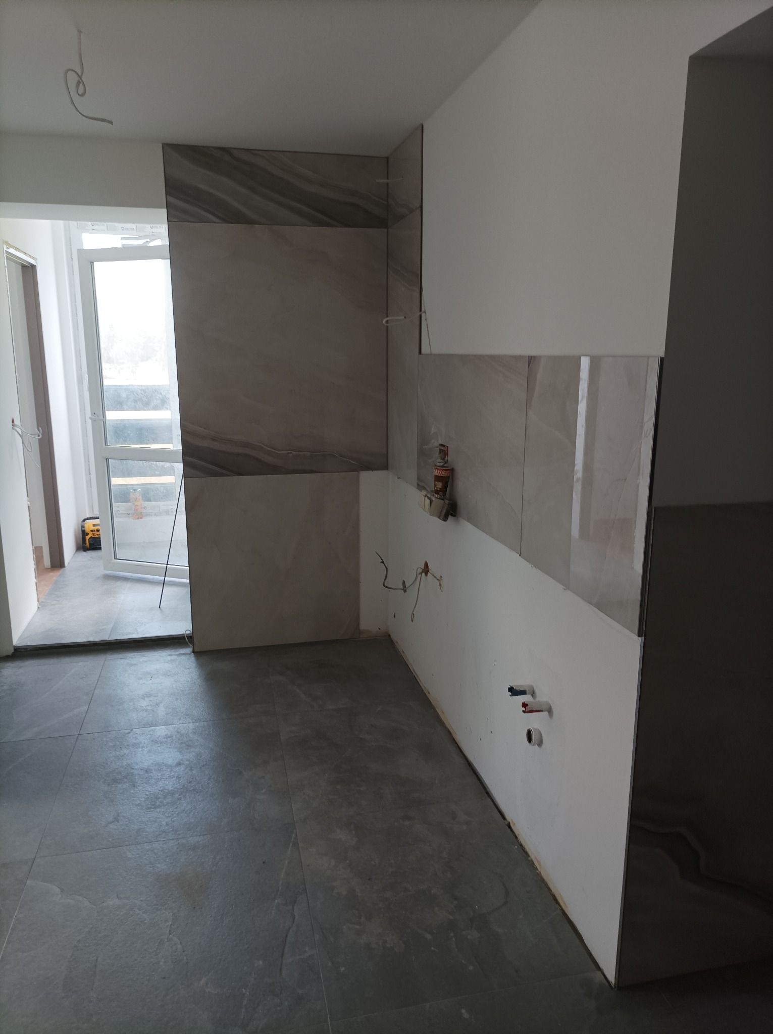 Wnętrze kuchni w trakcie układania płytek ściennych w odcieniach szarości, widoczne niedokończone instalacje wodne i fragment okna z widokiem na zewnątrz.