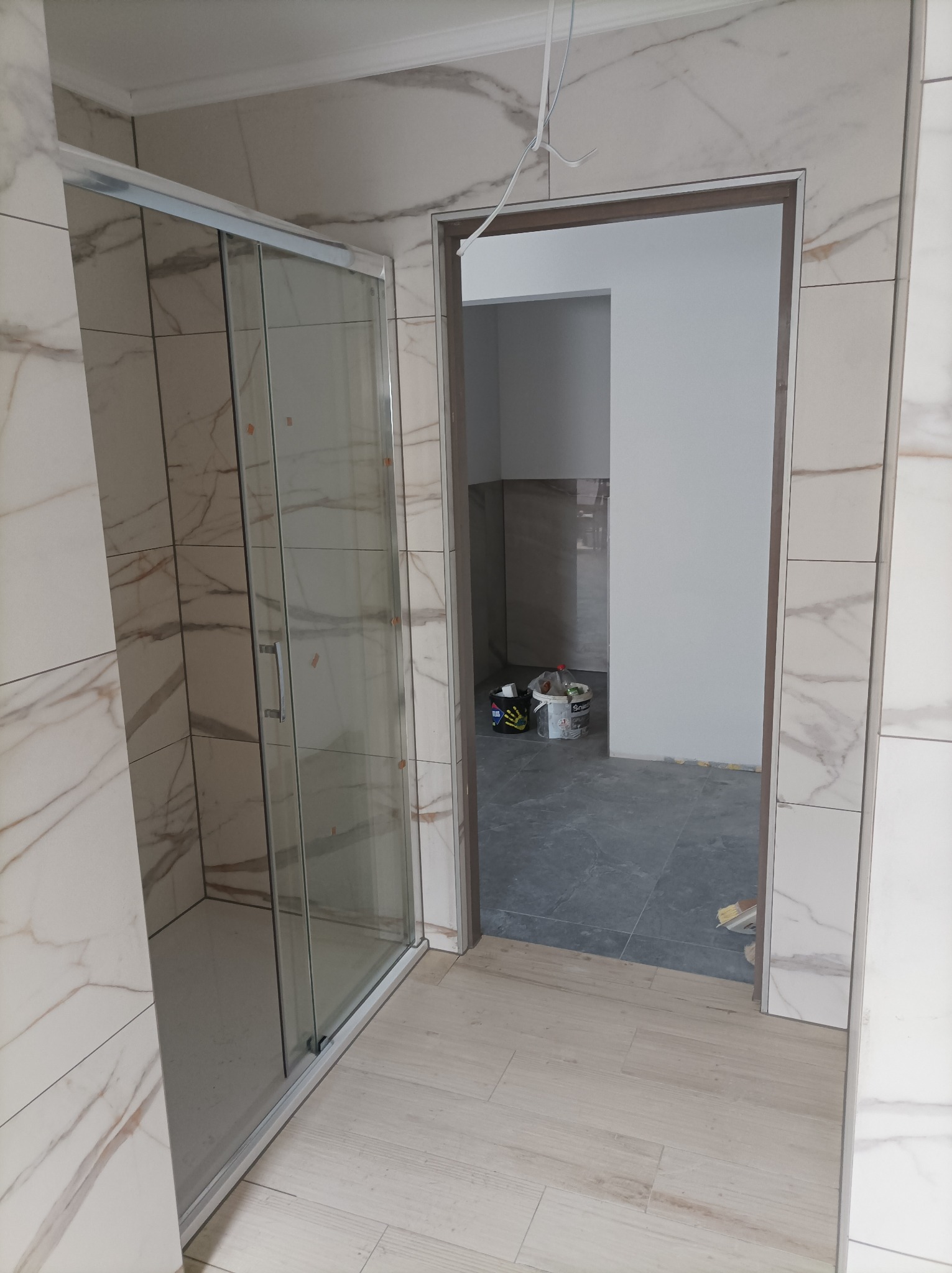 Wykończona łazienka z kabiną prysznicową i marmurowymi płytkami na ścianach, widok na sąsiednie pomieszczenie z szarymi płytkami podłogowymi i wiadrami po farbie.