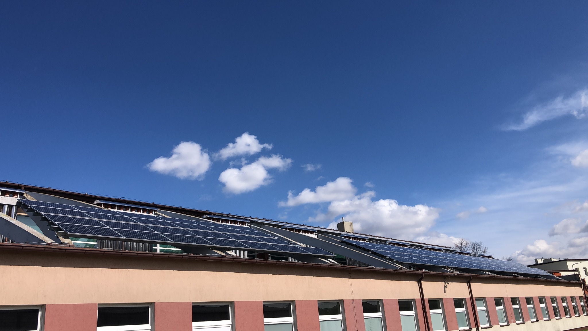 Rząd paneli słonecznych zamontowanych na dachu budynku z jasną fasadą i białymi oknami, na tle błękitnego nieba z kilkoma obłokami.