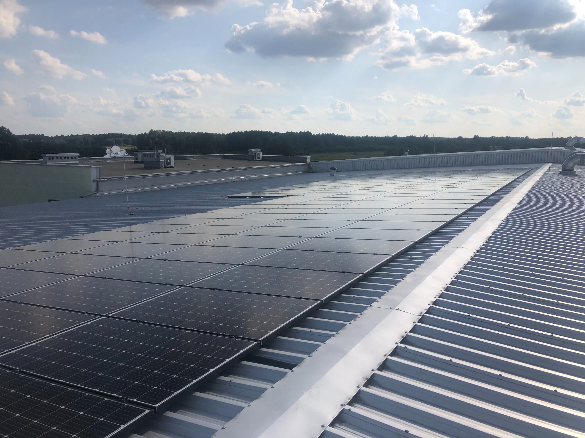 Panele słoneczne na blaszanym dachu budynku przemysłowego, widok z góry, błękitne niebo z chmurami w tle.