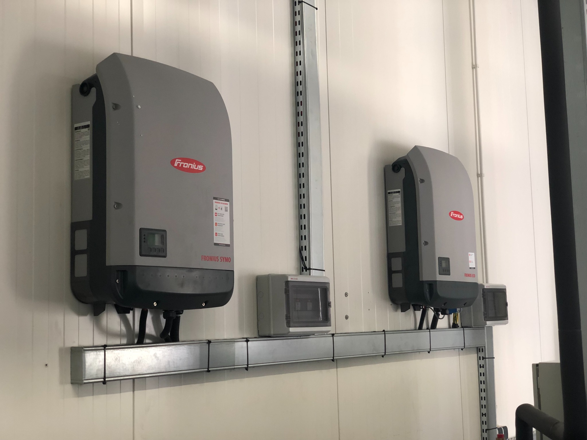 Dwa szare inwertery Fronius zamontowane na metalowej szynie na białej ścianie z widoczną skrzynką rozdzielczą i pionową listwą montażową.