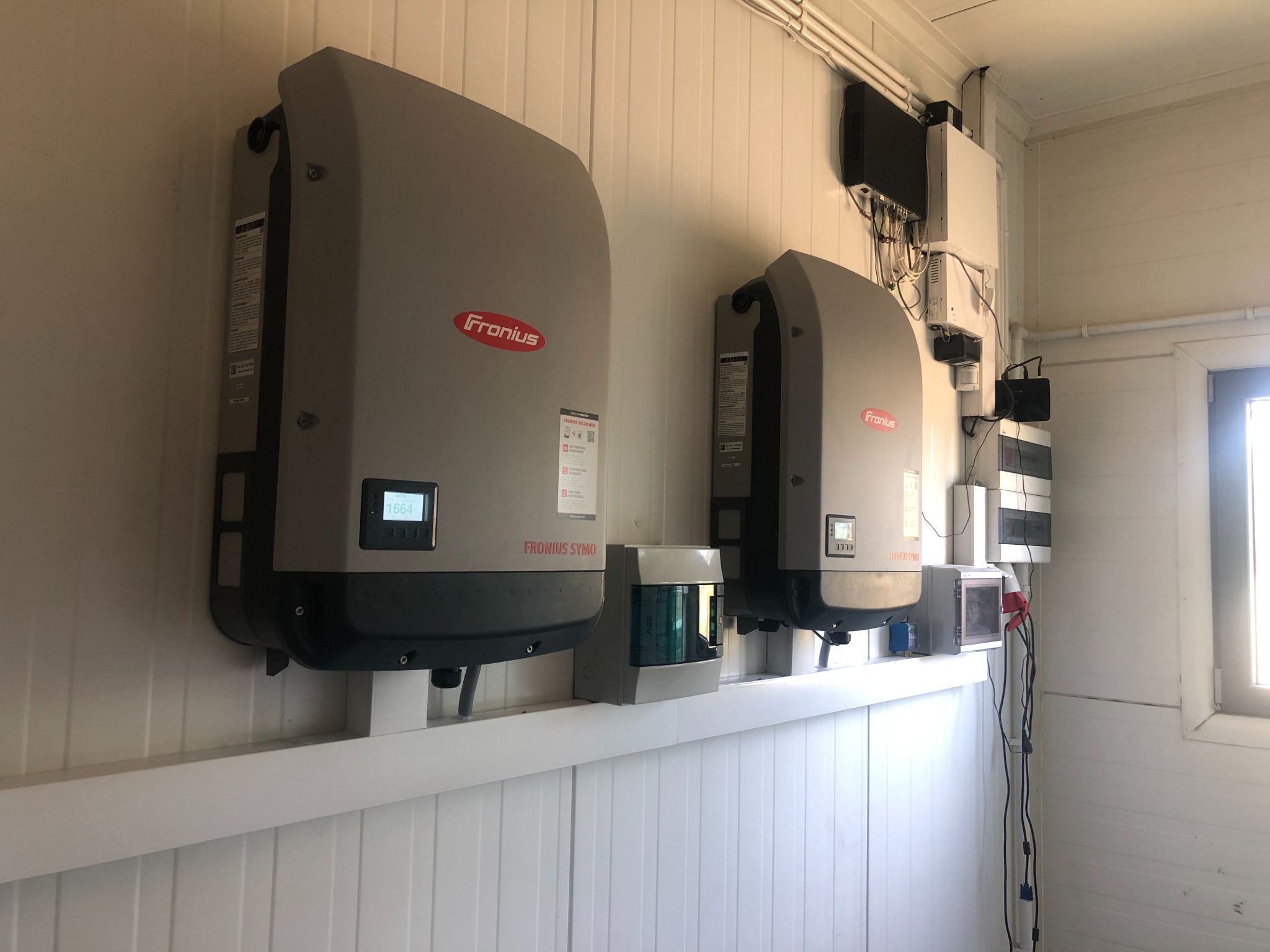 Dwa szare inwertery Fronius Symo zamontowane na białej ścianie z paneli w Krasocinie, widoczne okablowanie i dodatkowe urządzenia elektryczne.