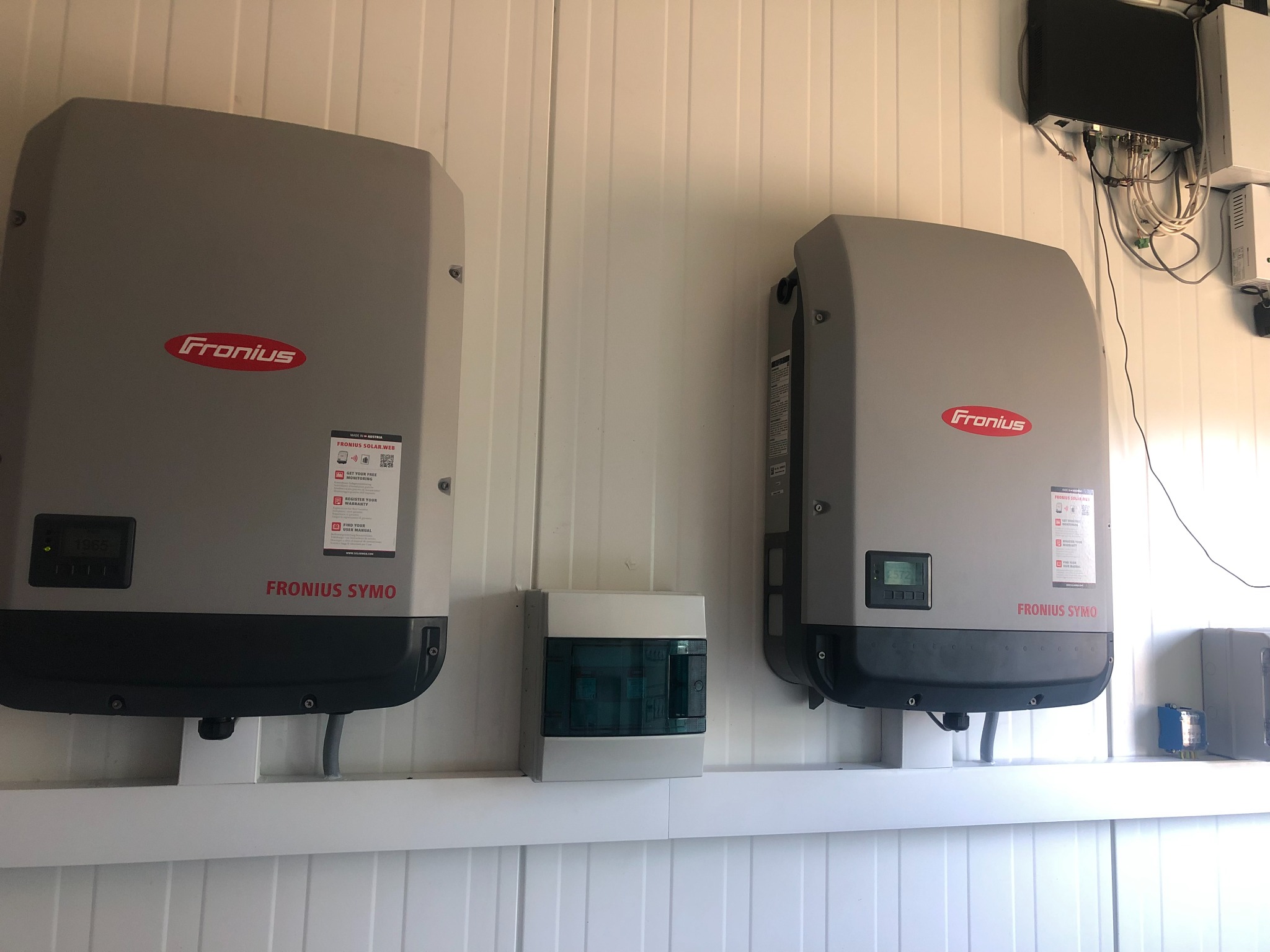 Dwa falowniki Fronius Symo zamontowane na białej ścianie z paneli, obok skrzynka zabezpieczeń, widoczne okablowanie i dodatkowe urządzenia elektryczne.