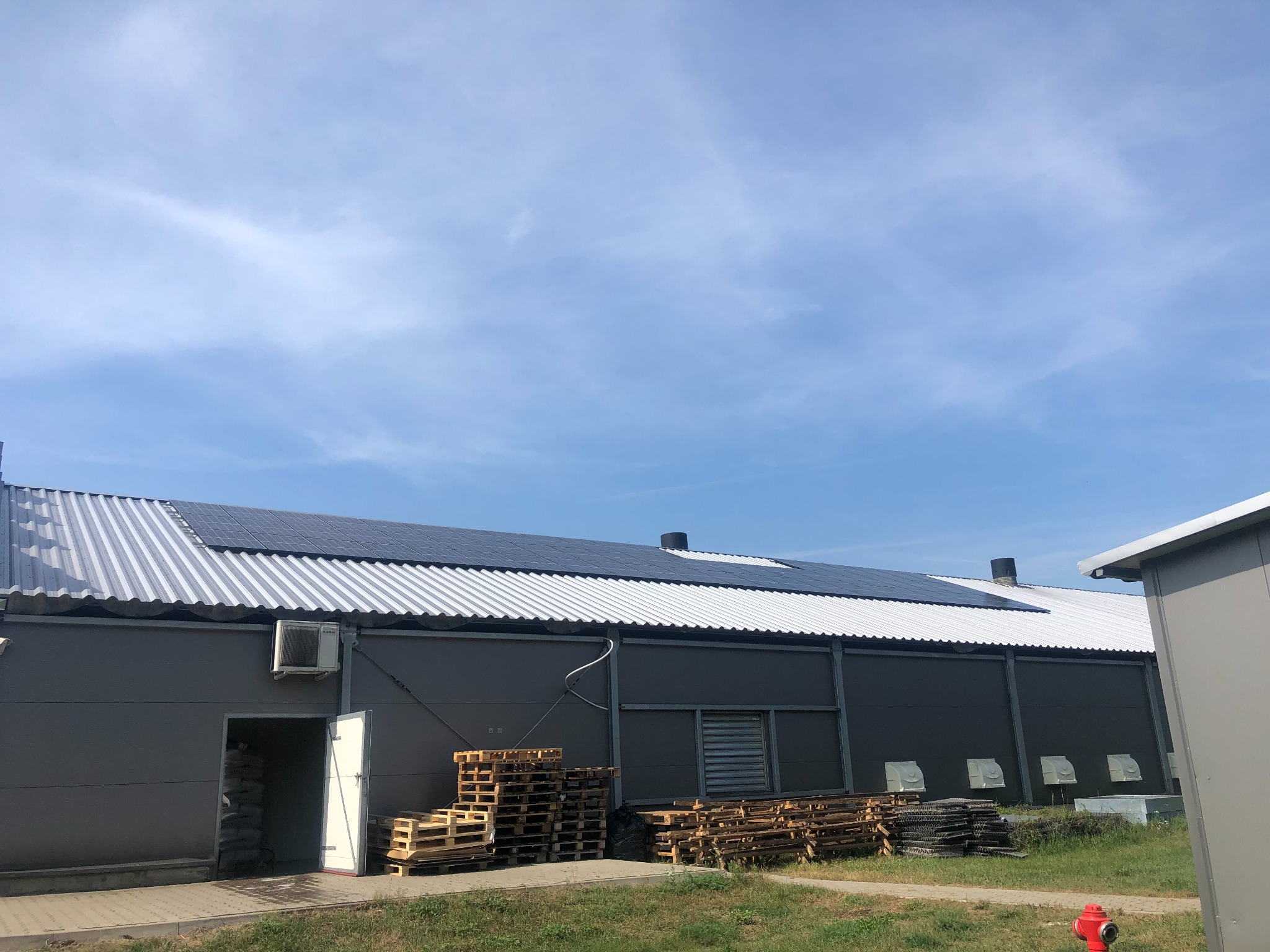 Instalacja paneli słonecznych na dachu hali przemysłowej, widoczne palety drewniane i hydrant przeciwpożarowy na trawniku.