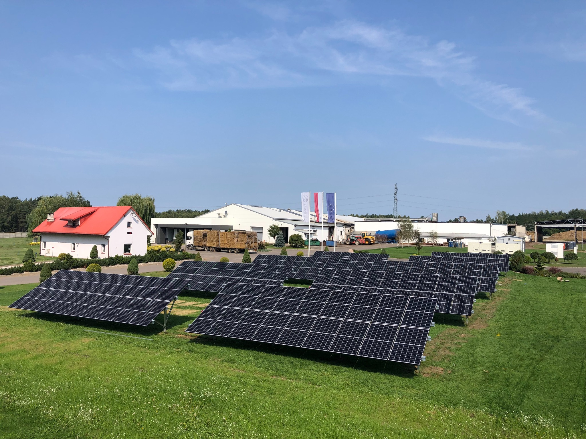 Rząd paneli słonecznych na trawiastym terenie, w tle budynki gospodarcze i dom z czerwonym dachem w słoneczny dzień.