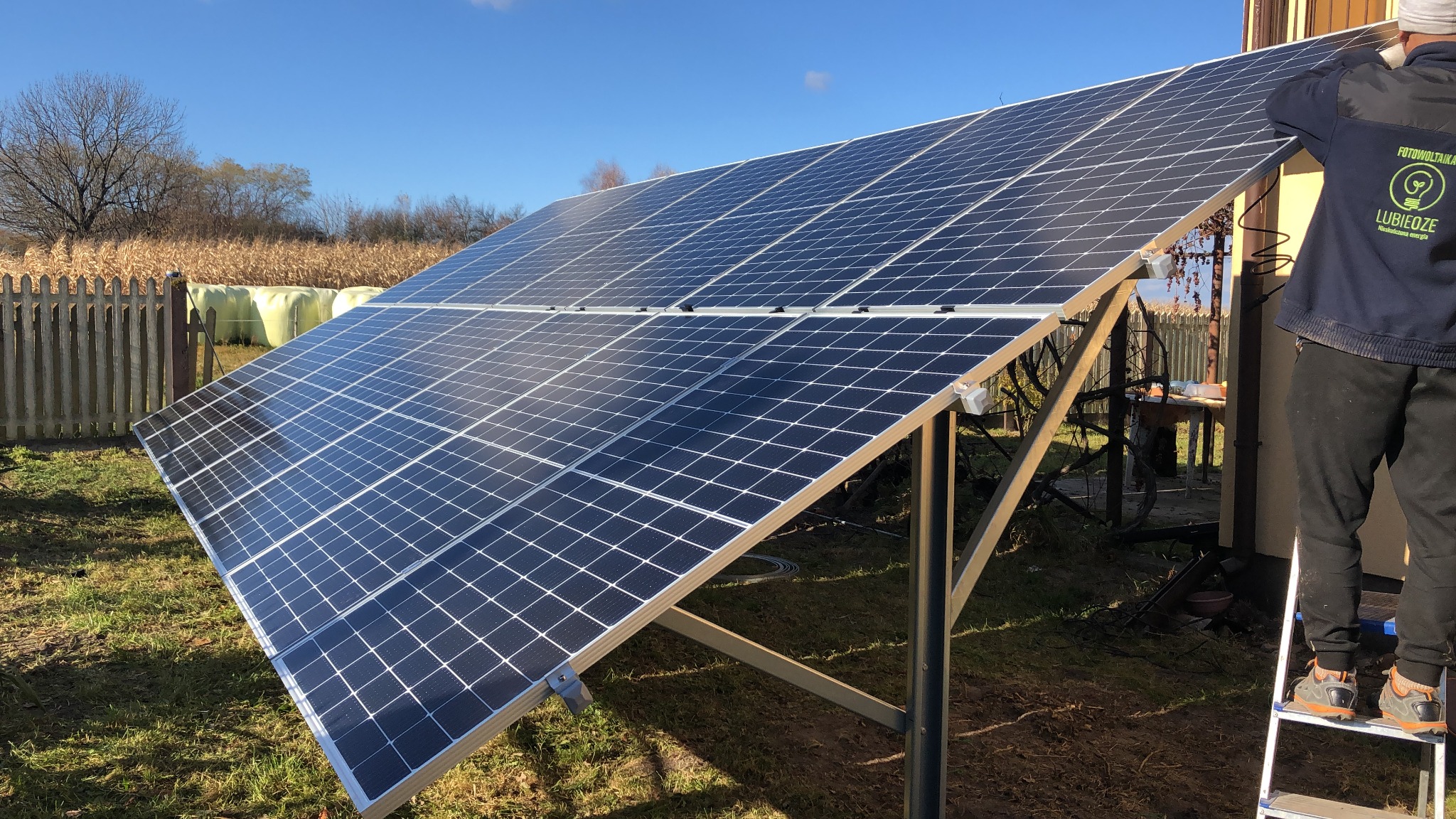 Montaż paneli fotowoltaicznych na konstrukcji wsporczej przy budynku, widoczny instalator na drabinie i logo firmy na bluzie, w tle ogrodzenie i pole kukurydzy.