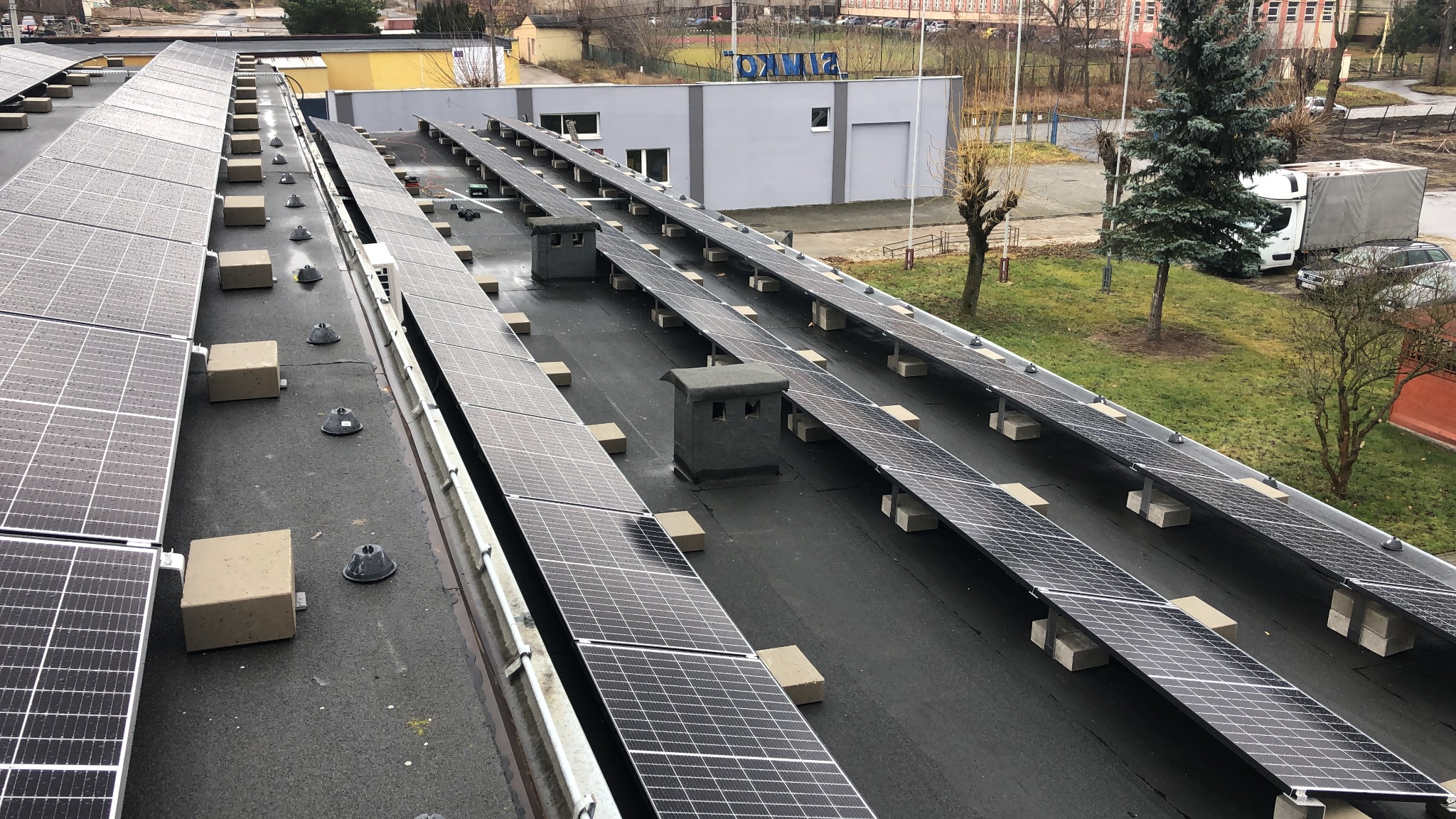 Rząd paneli słonecznych zamontowanych na płaskim dachu budynku przemysłowego, widoczne betonowe podstawy i elementy mocujące, w tle budynek z napisem 'SIMKO' i drzewa.