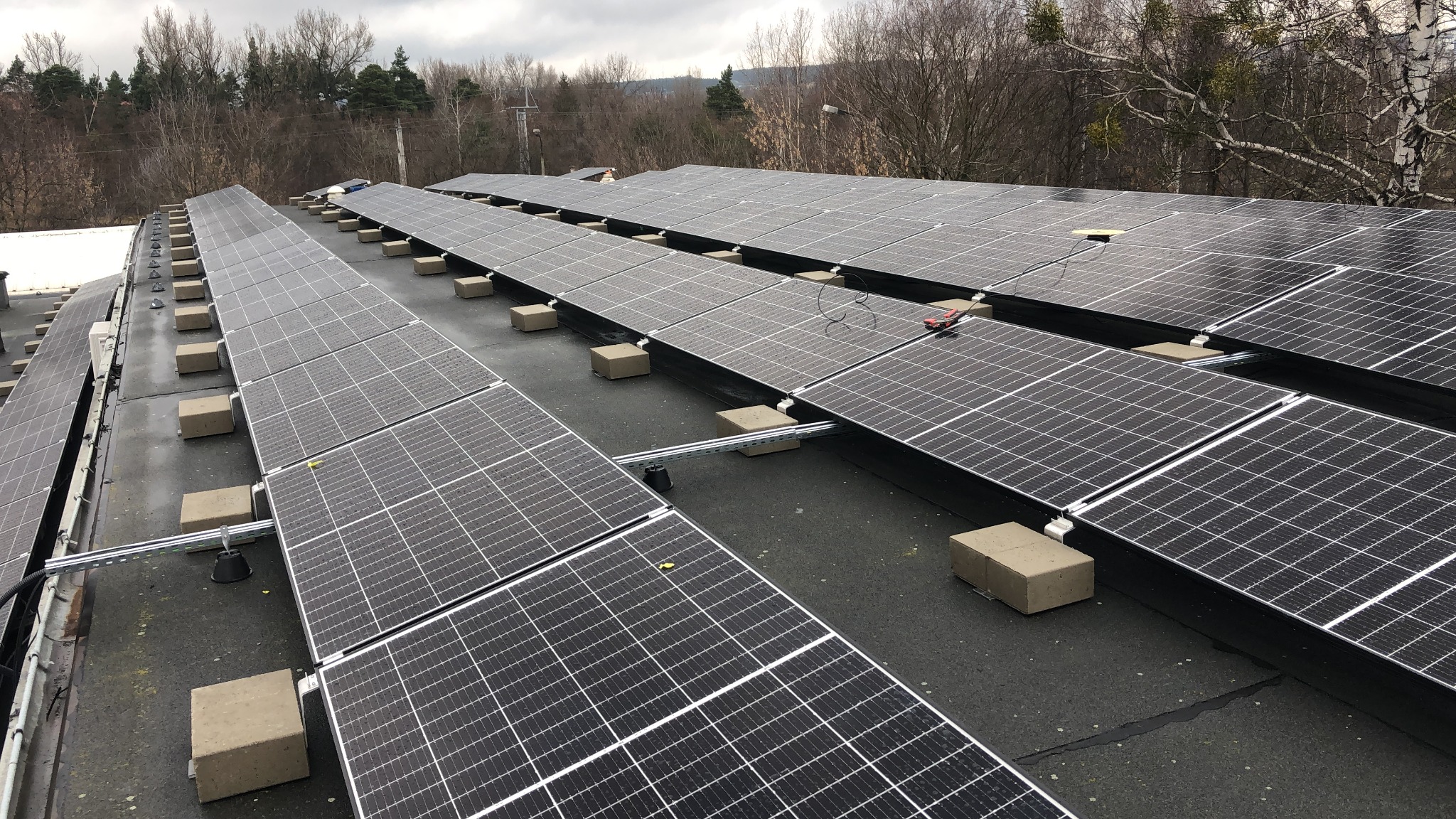 Rząd paneli słonecznych zamontowanych na płaskim dachu, z widocznymi elementami mocującymi i narzędziami na jednym z paneli, w tle drzewa bez liści.