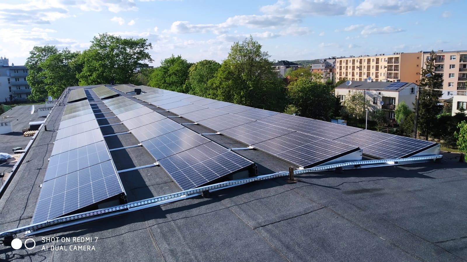 Rząd paneli słonecznych na płaskim dachu budynku, widok z góry, szyny montażowe, budynki mieszkalne w tle.