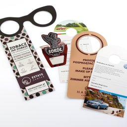 ANS Group Spółka z ograniczona odpowidzialnością - Zestaw zawieszek na klamkę o różnych wzorach i kolorach, prezentujących oferty firm z branży nieruchomości, gastronomii i motoryzacji na białym tle.