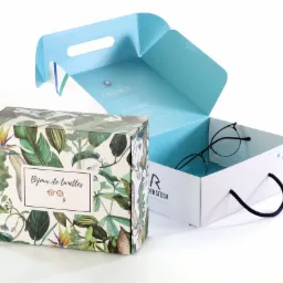 Dwa pudełka: jedno z motywem roślinnym i napisem, drugie białe z uchwytem, otwarte, z okularami w środku. Opakowanie firmowe z logo.