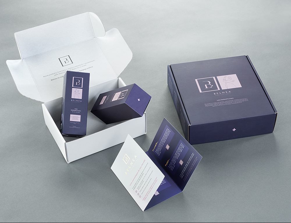 Zestaw kosmetyków marki Belnea w eleganckich, fioletowo-białych opakowaniach kartonowych, z ulotką informacyjną, na gładkim, szarym tle.