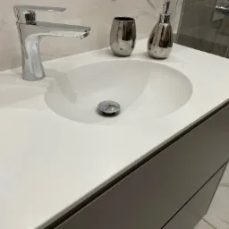 Nowoczesna, biała umywalka wpuszczana w szary blat z szafką, chromowana bateria i akcesoria łazienkowe na tle marmurowej ściany i kabiny prysznicowej.