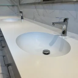 Biały blat łazienkowy z dwoma owalnymi umywalkami i chromowanymi bateriami, marmurowe płytki na ścianie w tle, widoczne uchwyty szafek pod blatem.