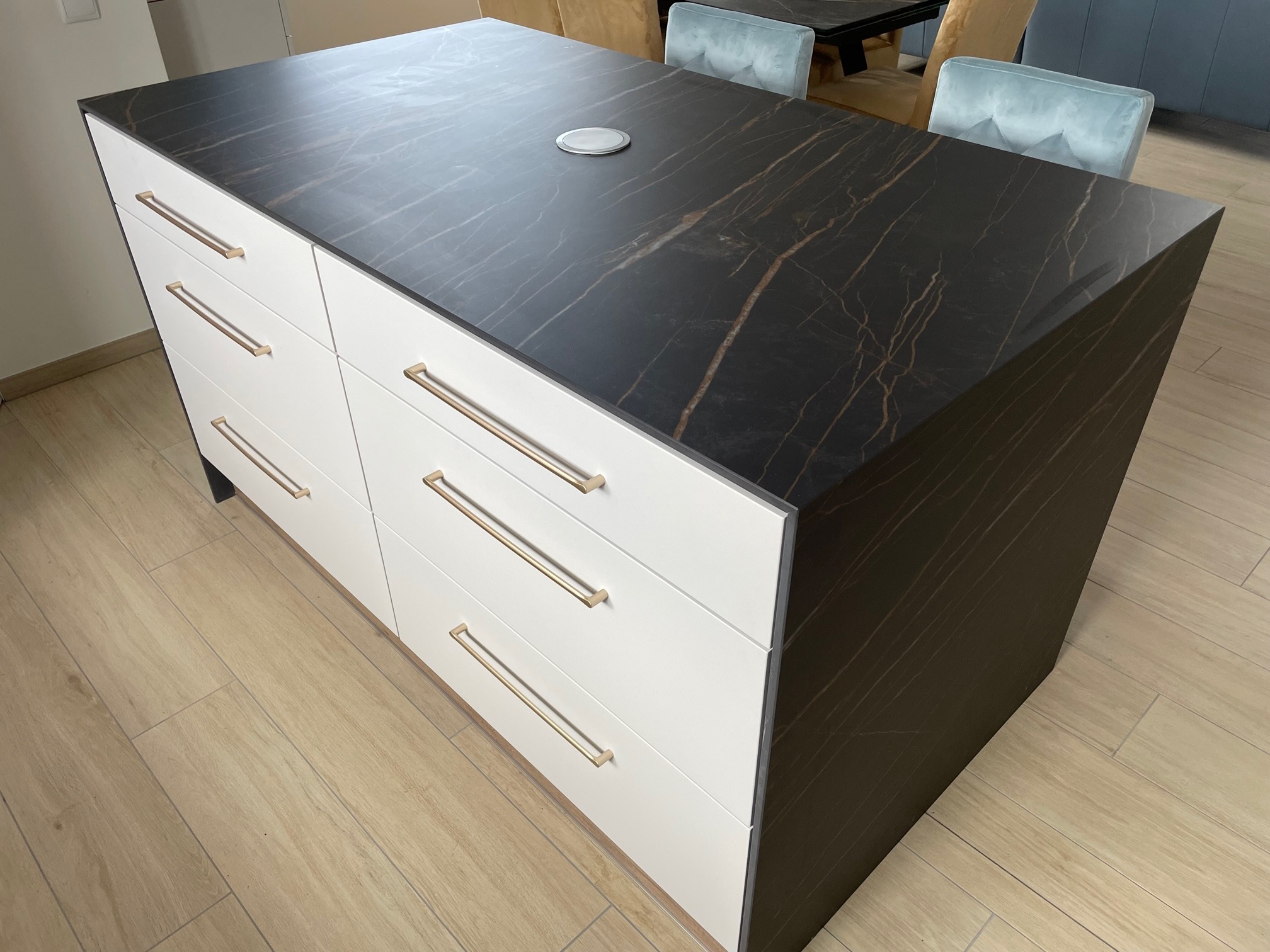 Wyspa kuchenna z białymi frontami szuflad, złotymi uchwytami i blatem imitującym czarny marmur z jasnymi żyłami, na drewnianej podłodze.