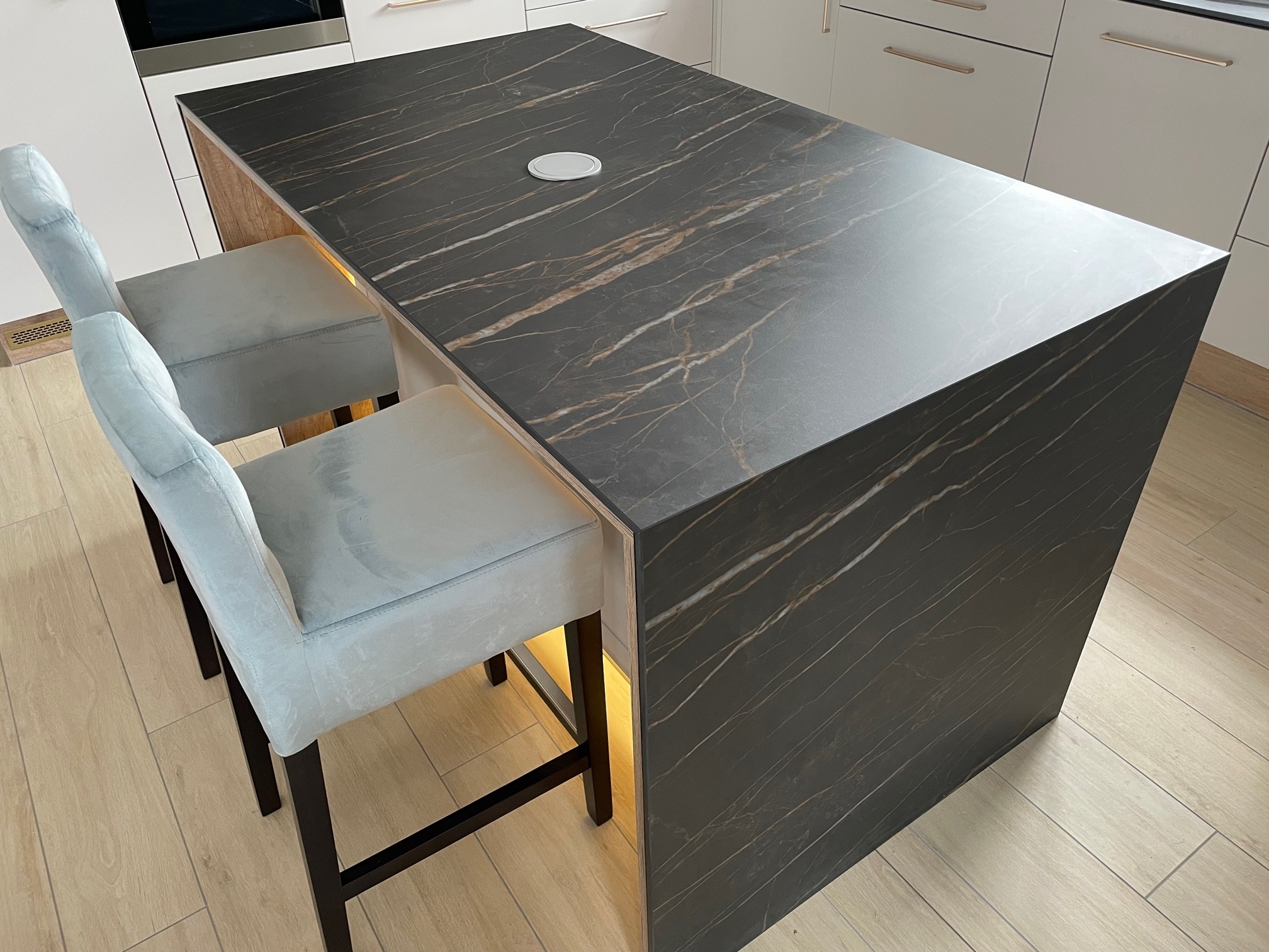 Wyspa kuchenna z blatem imitującym ciemny marmur z jasnymi żyłami, podświetlona od dołu, z dwoma krzesłami barowymi w kolorze jasnego popielu.