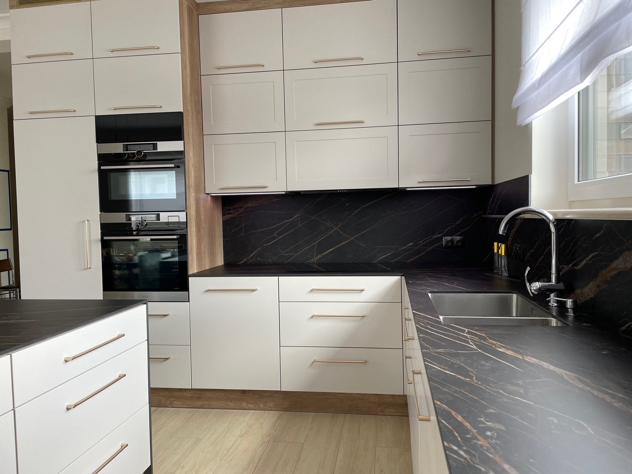 Nowoczesna kuchnia z białymi frontami szafek, złotymi uchwytami, blatem imitującym czarny marmur i wbudowanym piekarnikiem.