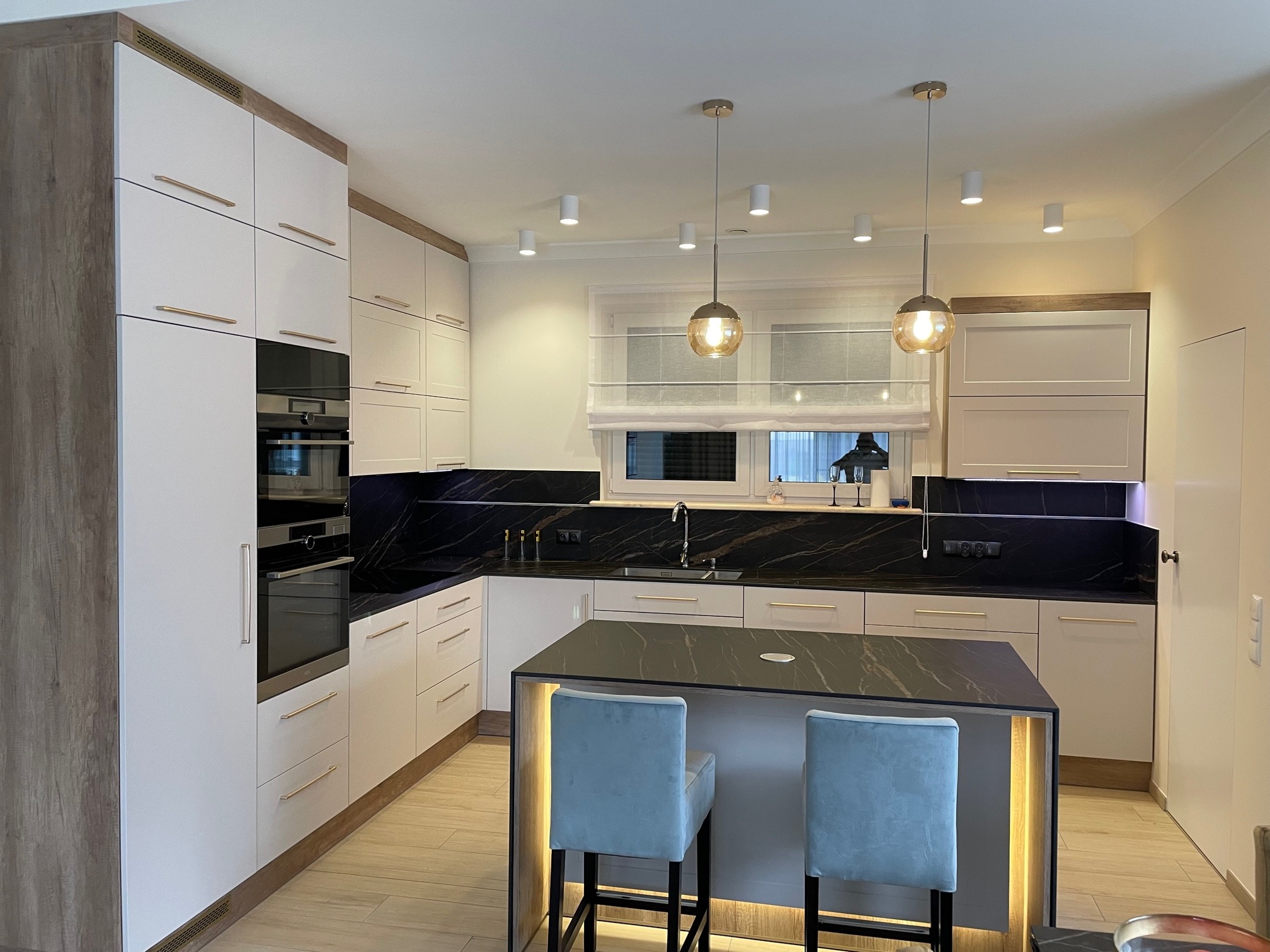 Nowoczesna kuchnia z białymi szafkami z drewnianymi akcentami, czarnym blatem i wyspą z podświetleniem LED oraz dwoma turkusowymi hokerami.