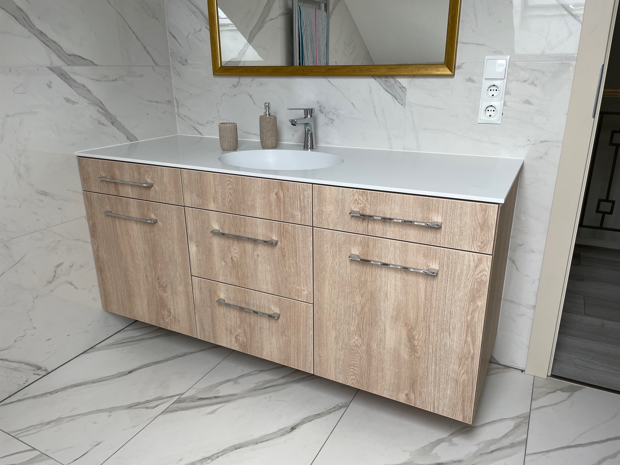 Nowoczesna szafka łazienkowa z drewnopodobnym frontem, białym blatem i owalną umywalką na tle marmurowej ściany i podłogi.