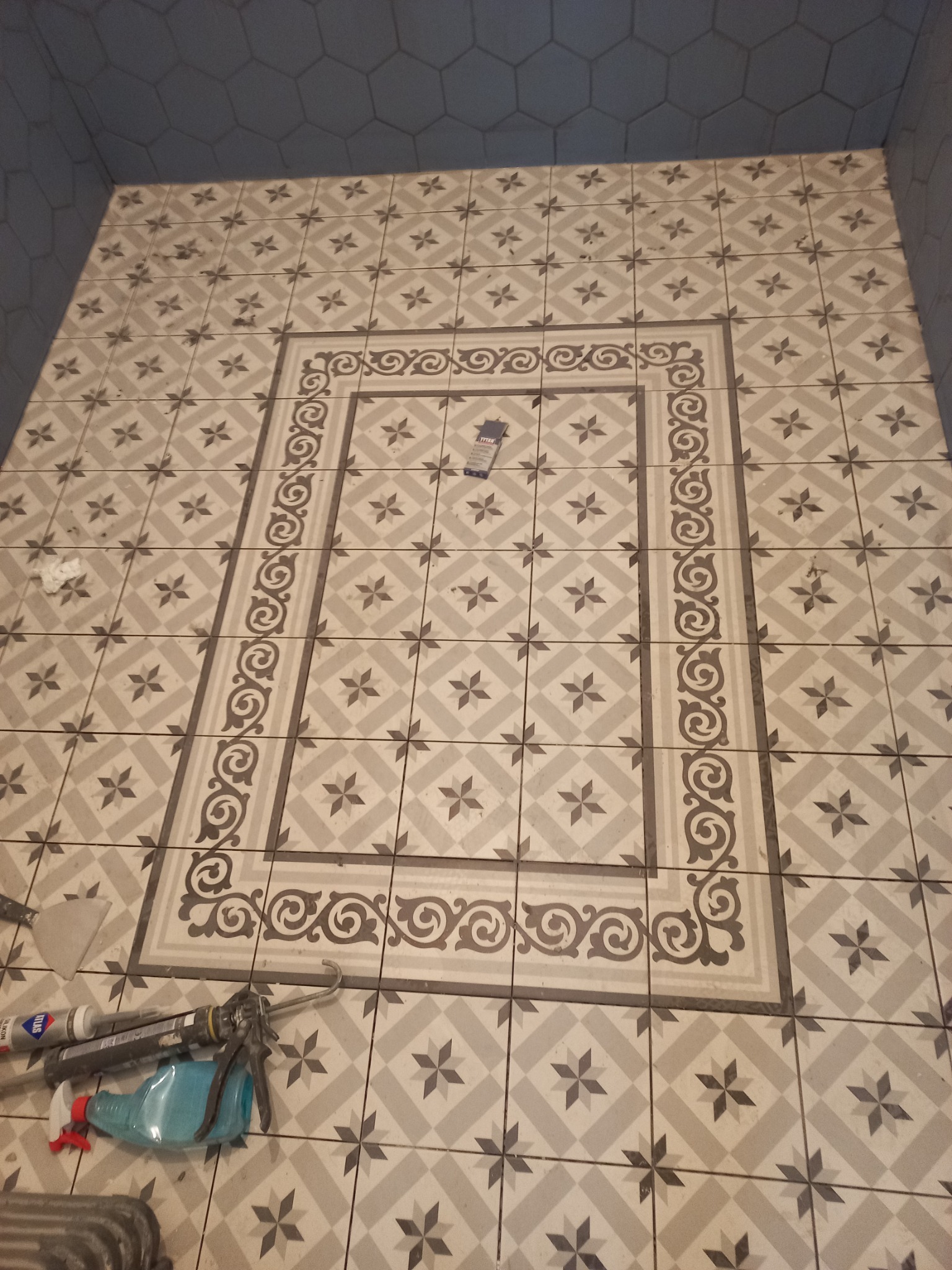 Ułożona podłoga w łazience z płytek w odcieniach szarości i beżu, z dekoracyjnym motywem w centralnej części, widoczne narzędzia glazurnicze i karton Tytan leżące na płytkach, ściany wykończone...