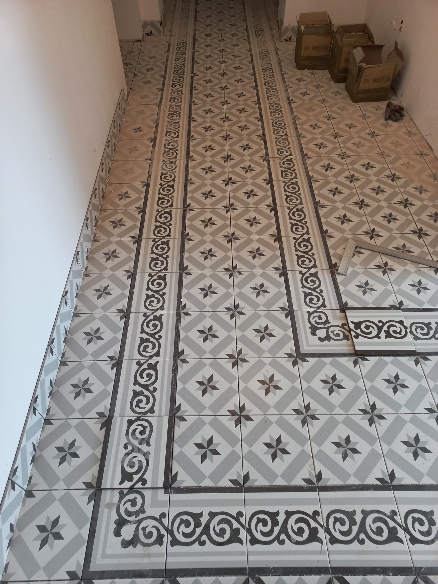 Ułożona podłoga z szaro-białych płytek z motywem gwiazd i dekoracyjnym pasem z ornamentami, widoczne kartony po płytkach i pojedyncze płytki do ułożenia.