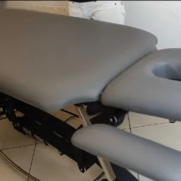 Szary, regulowany stół do zabiegów z otworem na twarz i podłokietnikami, widoczne metalowe elementy konstrukcyjne, w tle fragment osoby w białych spodniach.