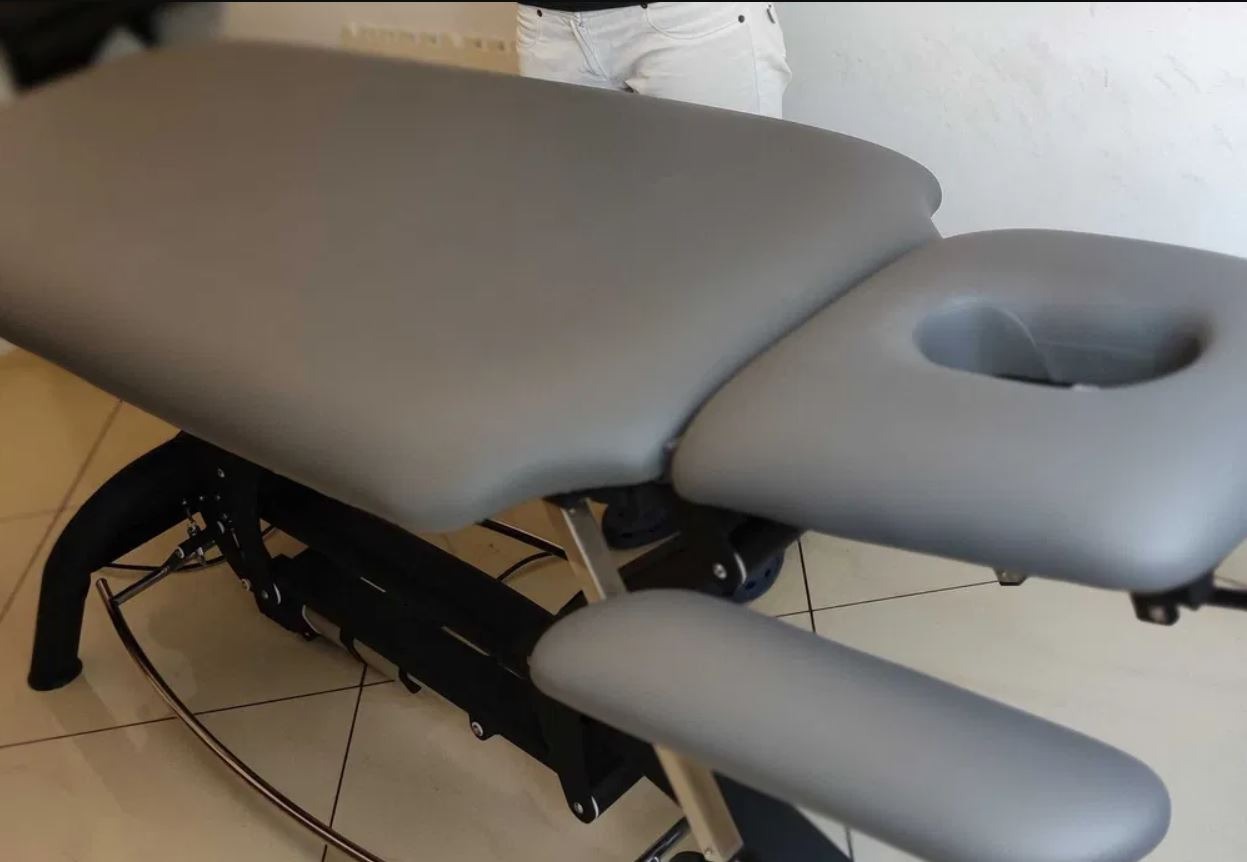 Szary, regulowany stół do zabiegów z otworem na twarz i podłokietnikami, widoczne metalowe elementy konstrukcyjne, w tle fragment osoby w białych spodniach.