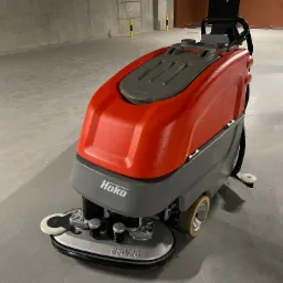Profesjonalna, samojezdna maszyna czyszcząca Hako 650/26 z czerwono-szarą obudową, widoczna na tle betonowej posadzki w hali garażowej, po której przejeździe widać mokry ślad.