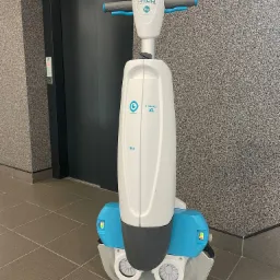 Profesjonalna, samojezdna maszyna czyszcząca I-Mop XL, z widocznymi szczotkami i niebieskimi zbiornikami na wodę, stoi na kafelkach w korytarzu biurowym z szarą ścianą o chropowatej fakturze...