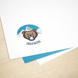Adrian Wiczyński - Stos arkuszy papieru z nadrukowanym logo firmy przedstawiającym niedźwiedzia na tle gór i hasłem 'Twoja Nazwa', leżący na drewnianym biurku.