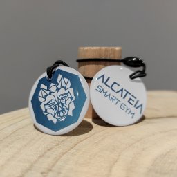 Adrian Wiczyński - Dwa okrągłe identyfikatory z logo i nazwą 'ALCATEIA SMART GYM', zawieszone na czarnych sznurkach, leżące na drewnianej powierzchni obok drewnianego walca z gumką.