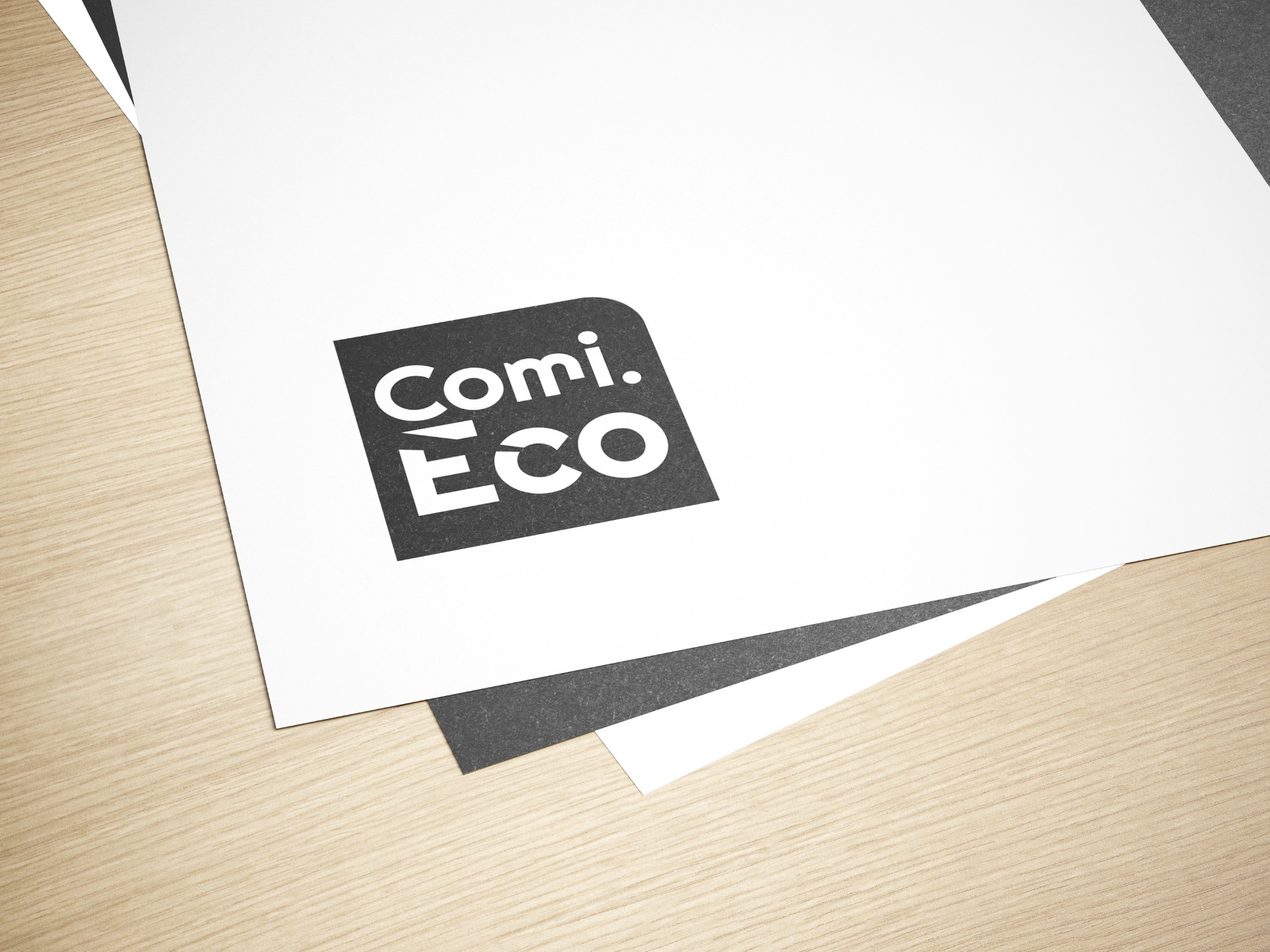 Stos kartek z nadrukowanym czarno-białym logo 'Comi. Eco' w lewym górnym rogu, ułożonych na jasnym drewnianym blacie.