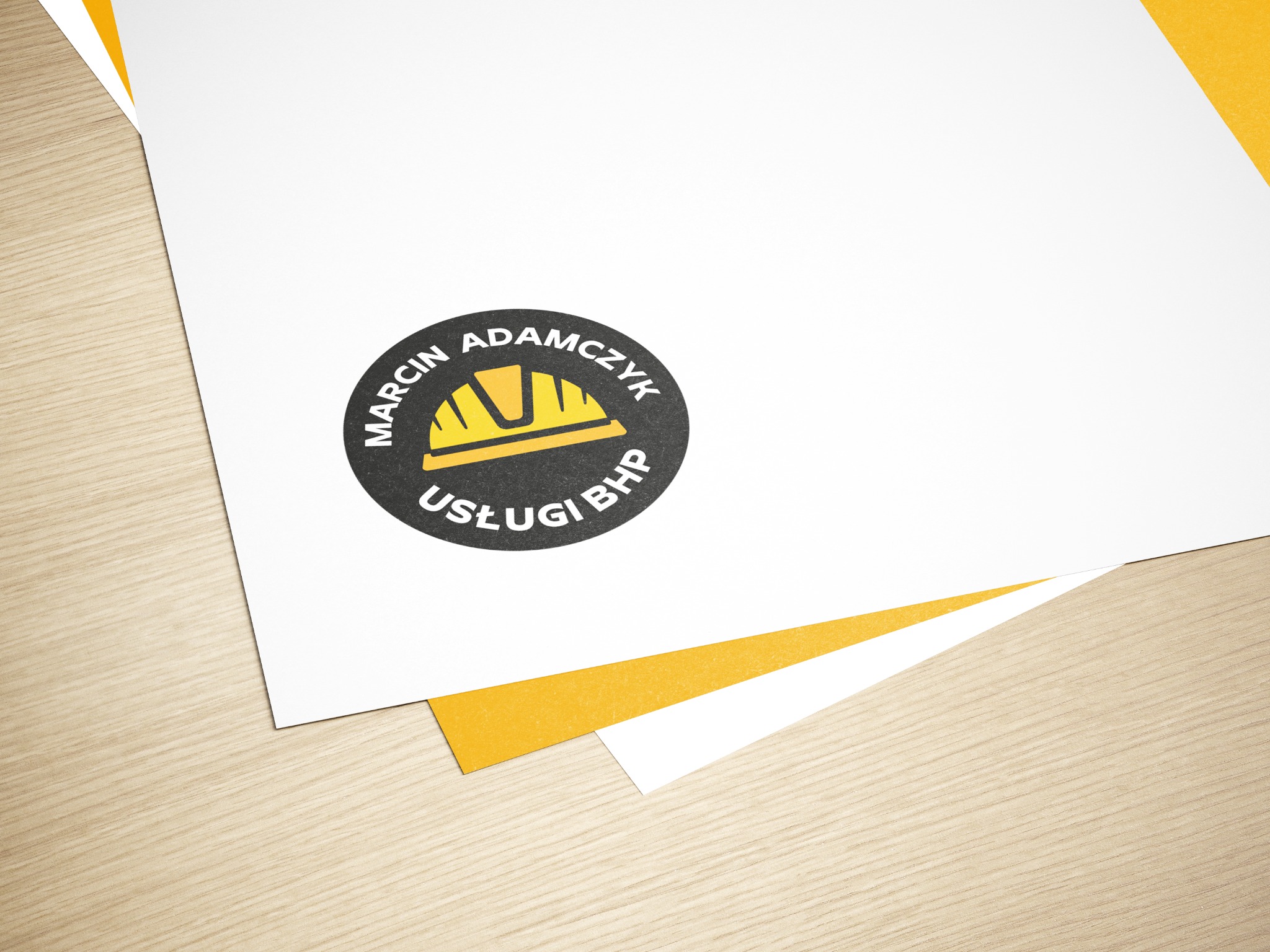 Wizytówka z logo firmy Marcin Adamczyk Usługi BHP przedstawiającym żółty kask na czarnym tle, leżąca na drewnianym stole.