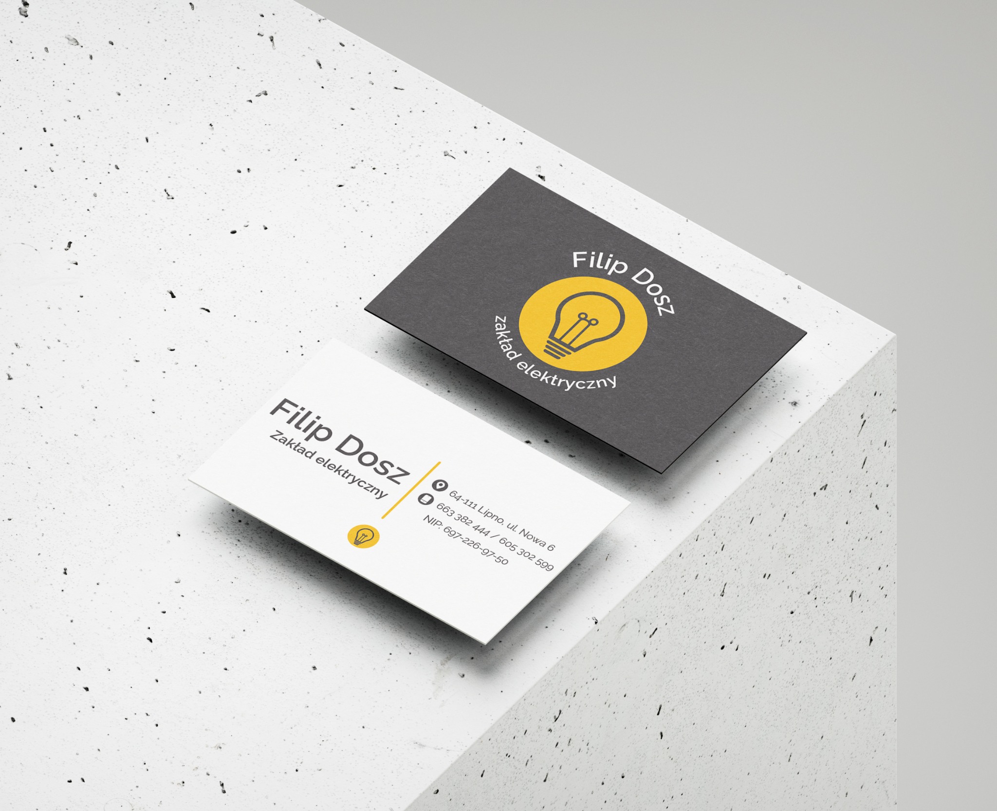 Dwie wizytówki na szarym, teksturowanym tle. Jedna biała z danymi kontaktowymi, druga grafitowa z logo firmy elektrycznej, żarówka w żółtym okręgu.