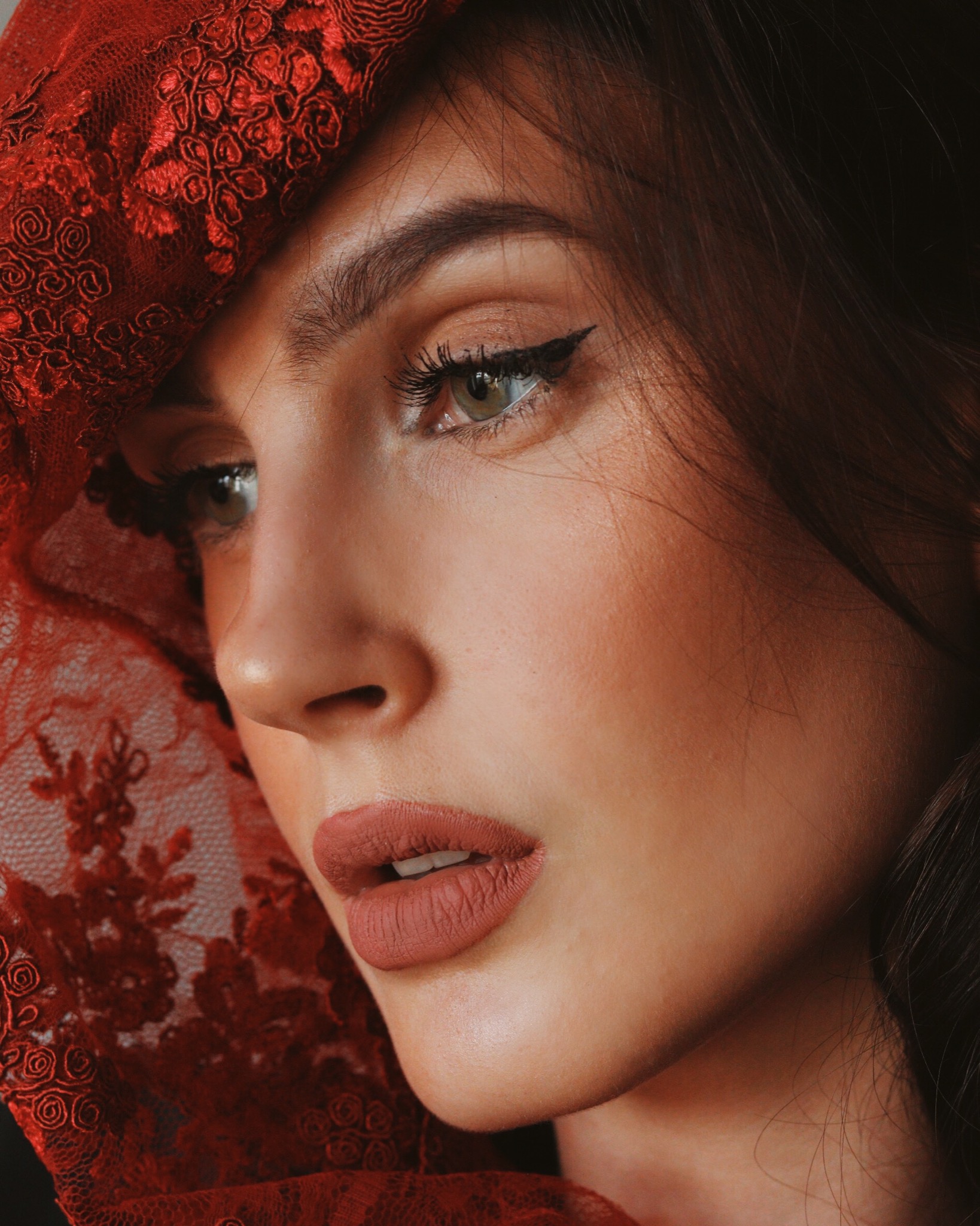 Portret kobiety z delikatnym makijażem, zarysowaną kreską na powiece i ustami w odcieniu nude, częściowo zasłoniętej czerwoną koronkową tkaniną.