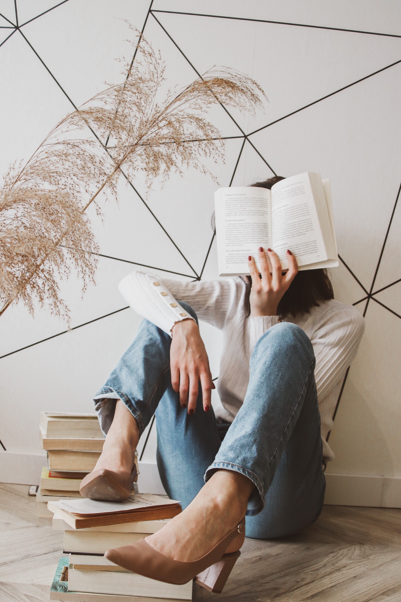 Kobieta siedząca na podłodze z książkami, zasłaniająca twarz otwartą książką, ubrana w dżinsy i białą bluzkę, w tle ściana z geometrycznym wzorem i pampasowa trawa.