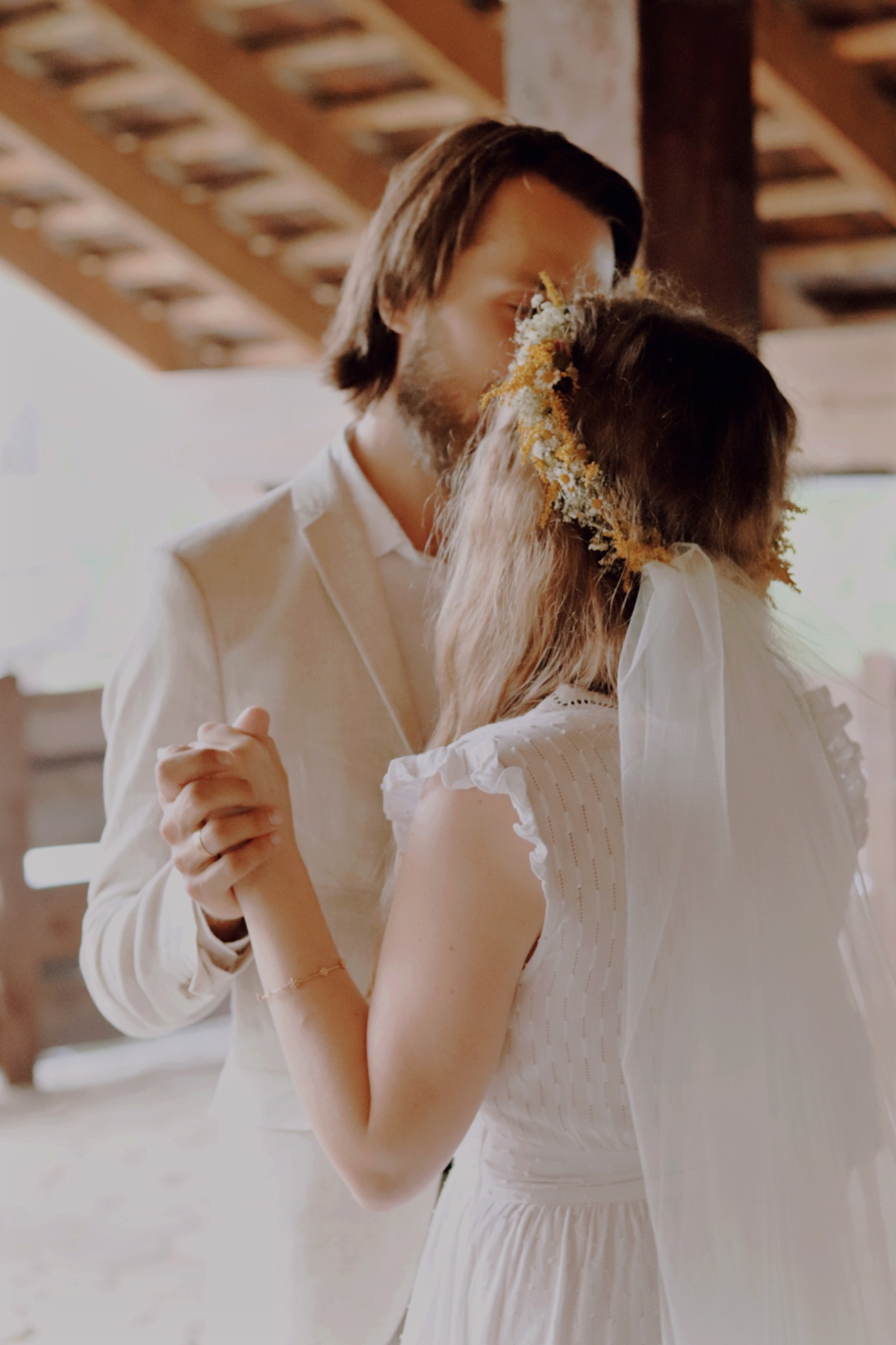 Para młoda trzymająca się za ręce podczas pierwszego tańca, panna młoda w białej sukni z koronkowymi detalami i wiankiem z polnych kwiatów, pan młody w kremowym garniturze, drewniana konstrukcja...