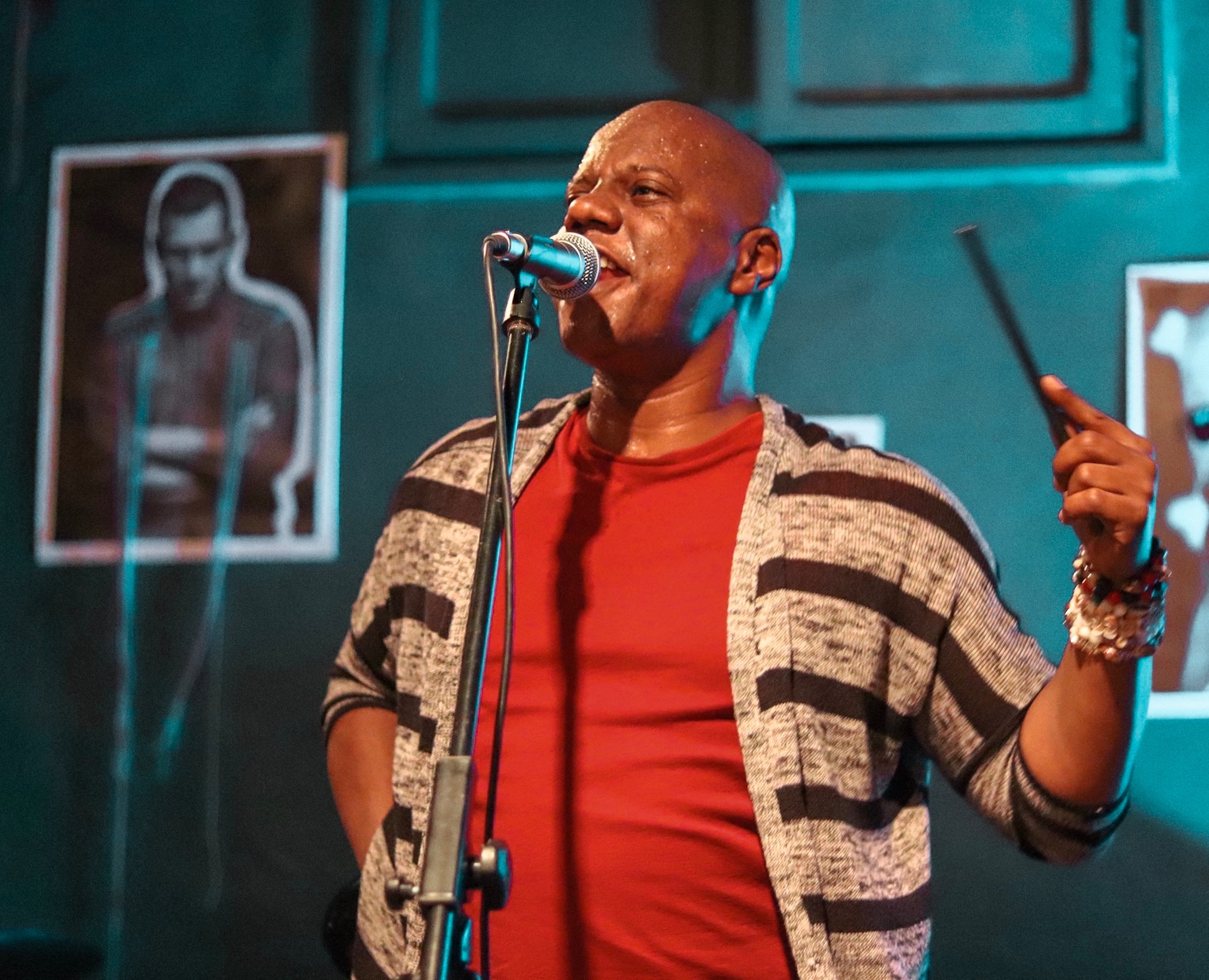 Wokalista z Wrocławia z mikrofonem śpiewa na scenie, trzymając pałeczkę perkusyjną w dłoni, w tle plakat z portretem.