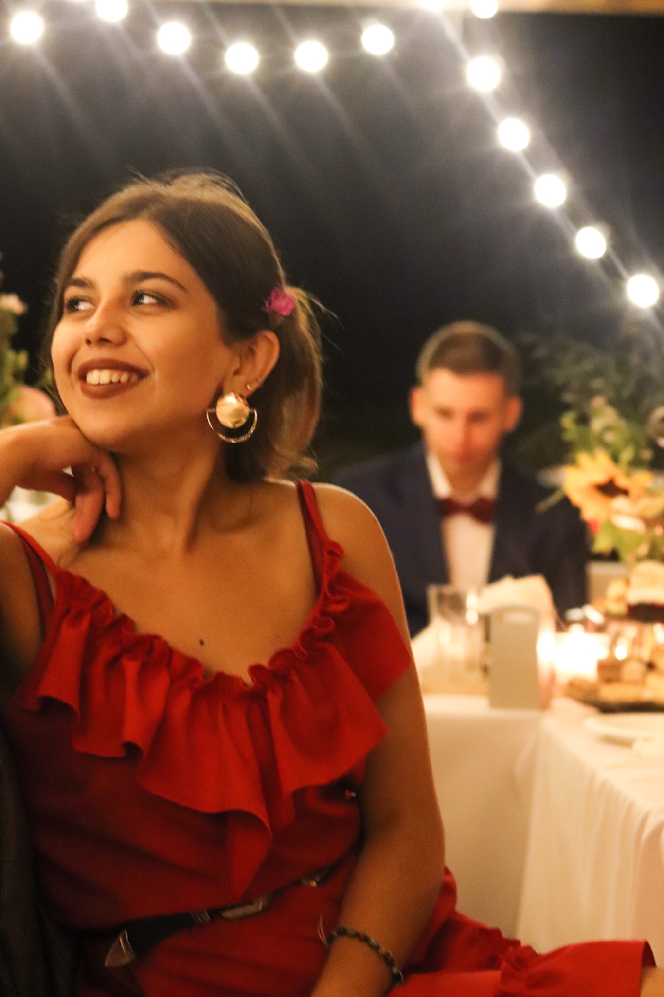 Uśmiechnięta kobieta w czerwonej sukience z falbankami i złotych kolczykach, siedząca przy stole udekorowanym kwiatami i świecami, z mężczyzną w tle podczas eventu oświetlonego girlandą żarówek.