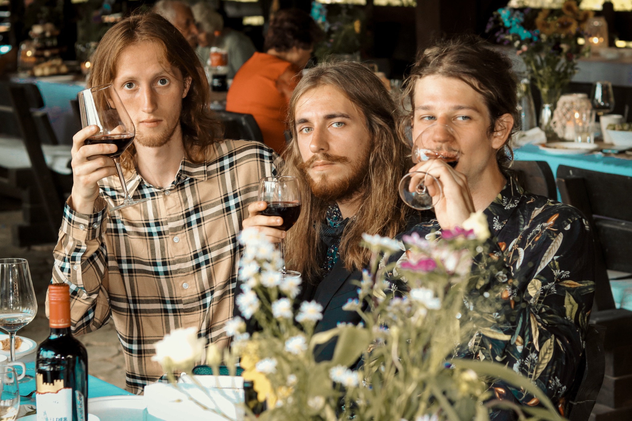 Trzech młodych mężczyzn z długimi włosami siedzi przy stole, trzymając kieliszki z czerwonym winem podczas wydarzenia plenerowego; widoczne kwiaty w wazonie na pierwszym planie oraz butelka wina.